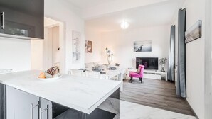 Une cuisine moderne avec un plan de travail blanc, un coin repas agrémenté d’une chaise rose et un téléviseur posé sur un meuble blanc.