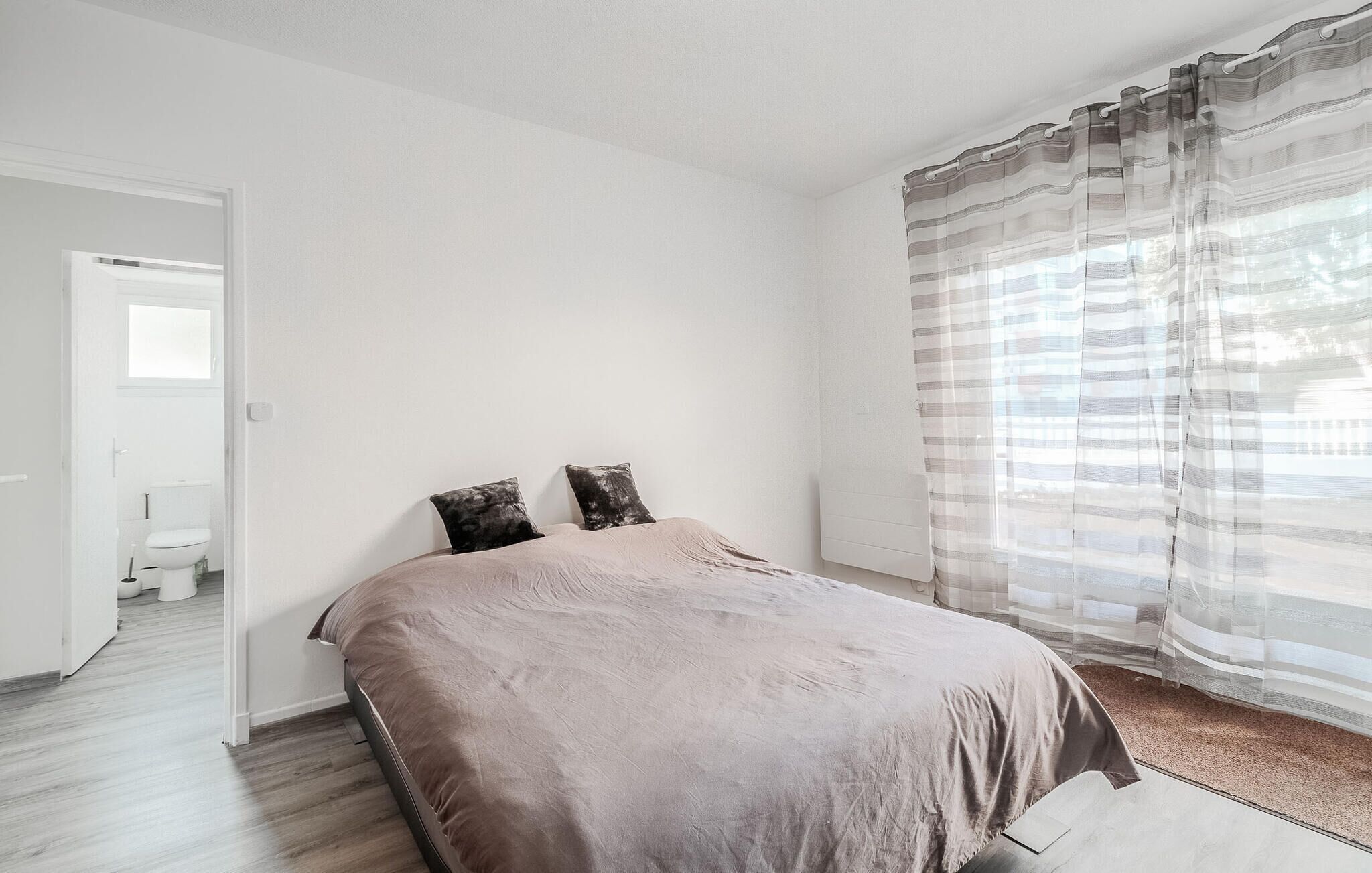 A bedroom with a bed, two pillows, a window with curtains, a door leading to a bathroom, and a wooden floor.