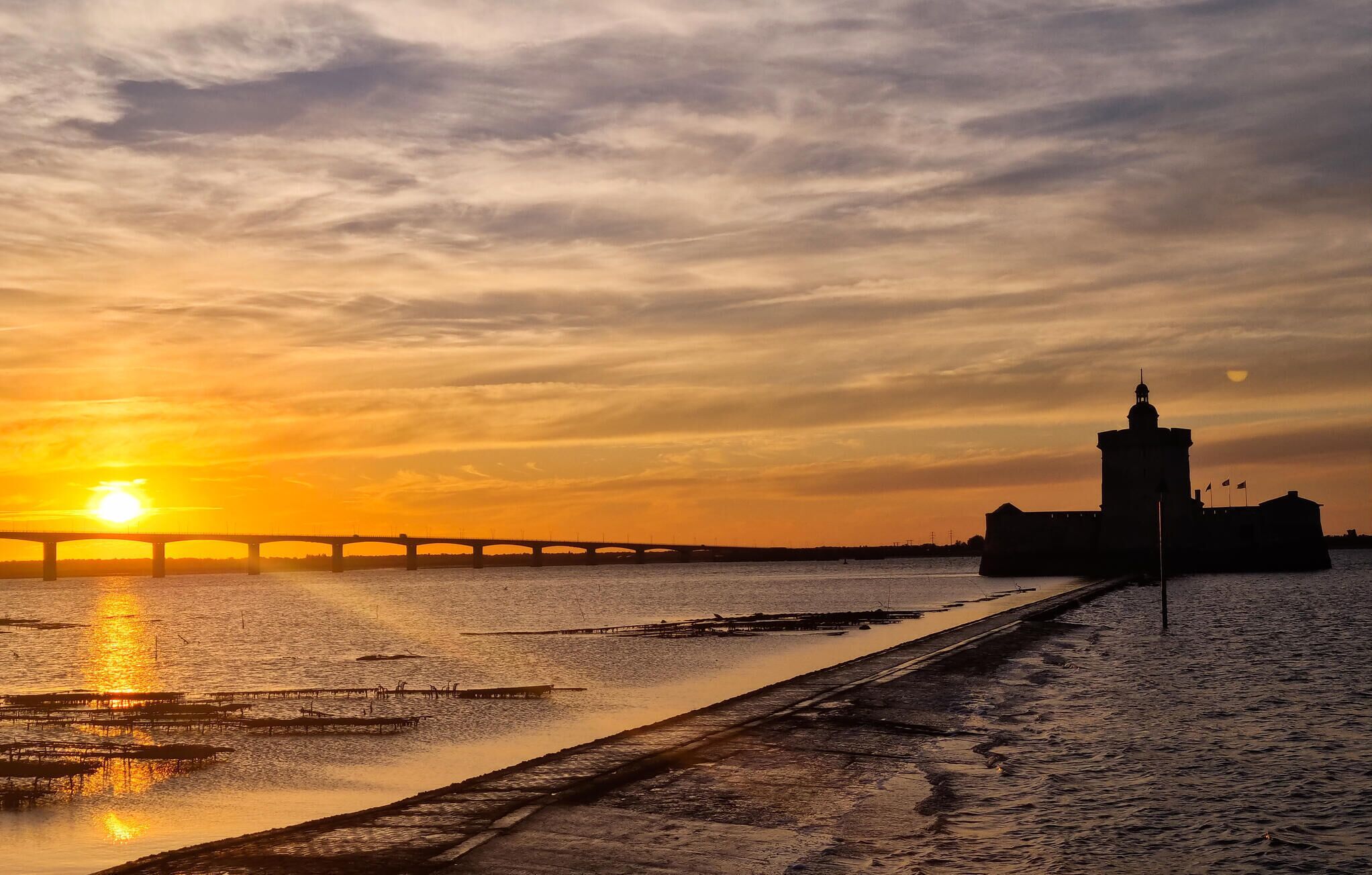 Solnedgang over en flod med en bro og en fæstningslignende bygning.