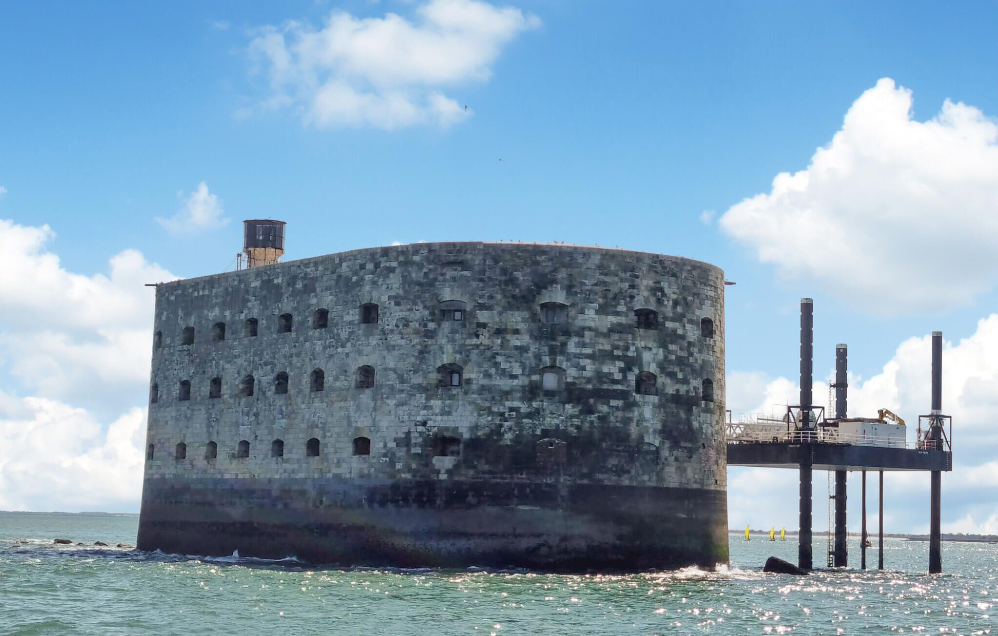 A large, cylindrical concrete structure with multiple small windows, situated in a body of water.