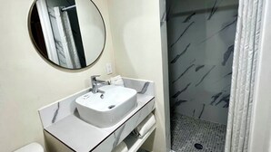 Une salle de bain moderne avec un sol en marbre, un miroir rond, un lavabo blanc et une douche dotée d’une porte en verre.