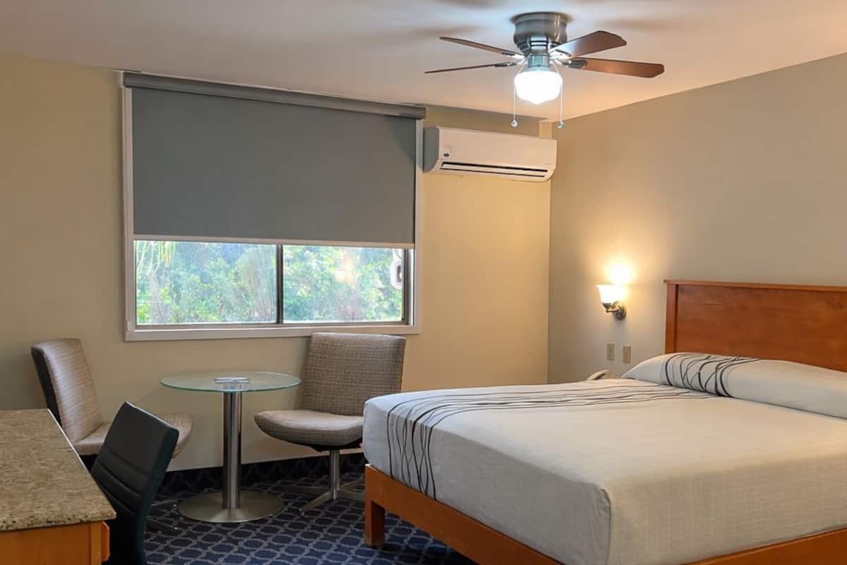 A hotel room with a bed, a desk, a chair, a small table, a ceiling fan, and an air conditioning unit.