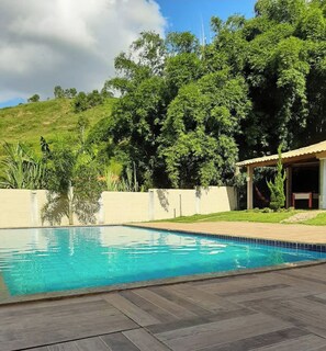 A swimming pool with a wooden deck, surrounded by greenery and a hill in the background. - Chácara do Allan (Manhuaçu)