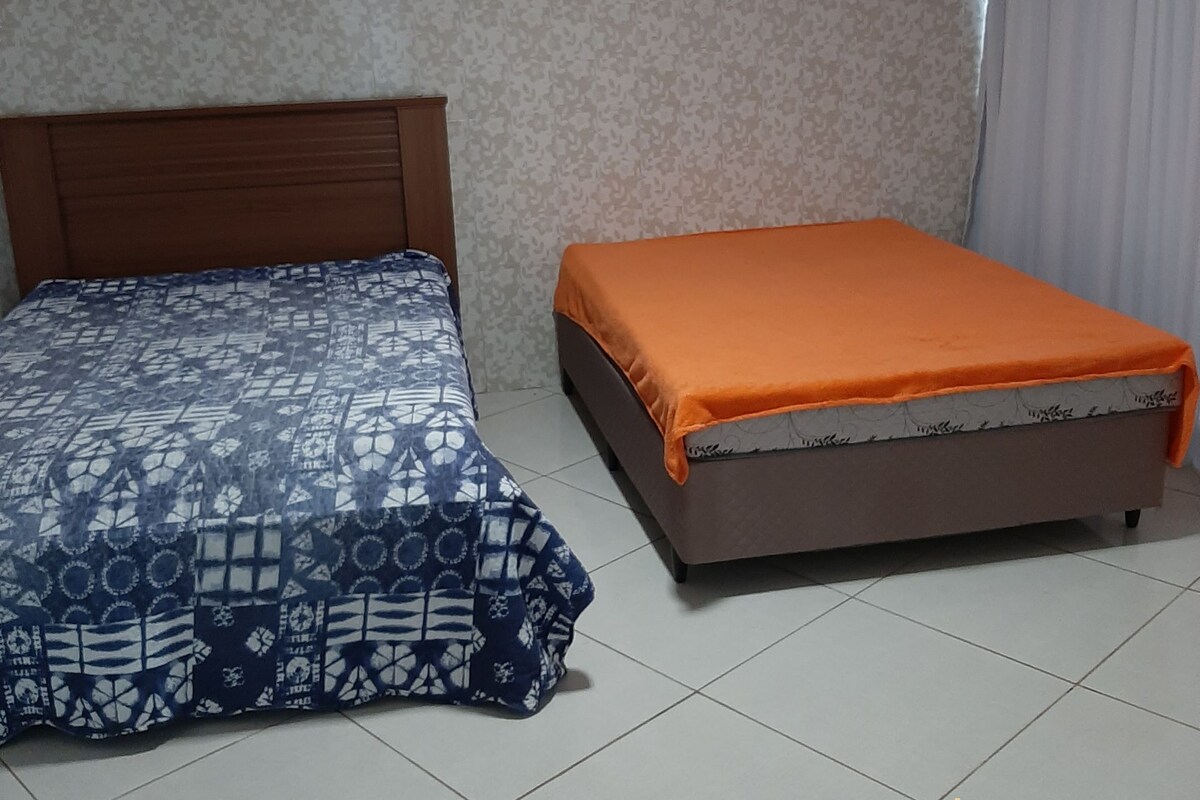 A room with two beds, one with a patterned bedspread and the other with an orange cover. The room has patterned wallpaper and a tiled floor.