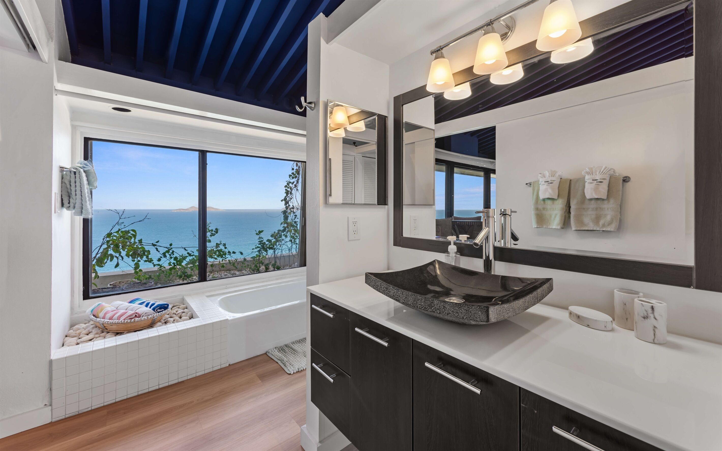 Une salle de bain moderne avec une grande fenêtre, un lavabo en pierre, des meubles sombres et une vue sur la mer.