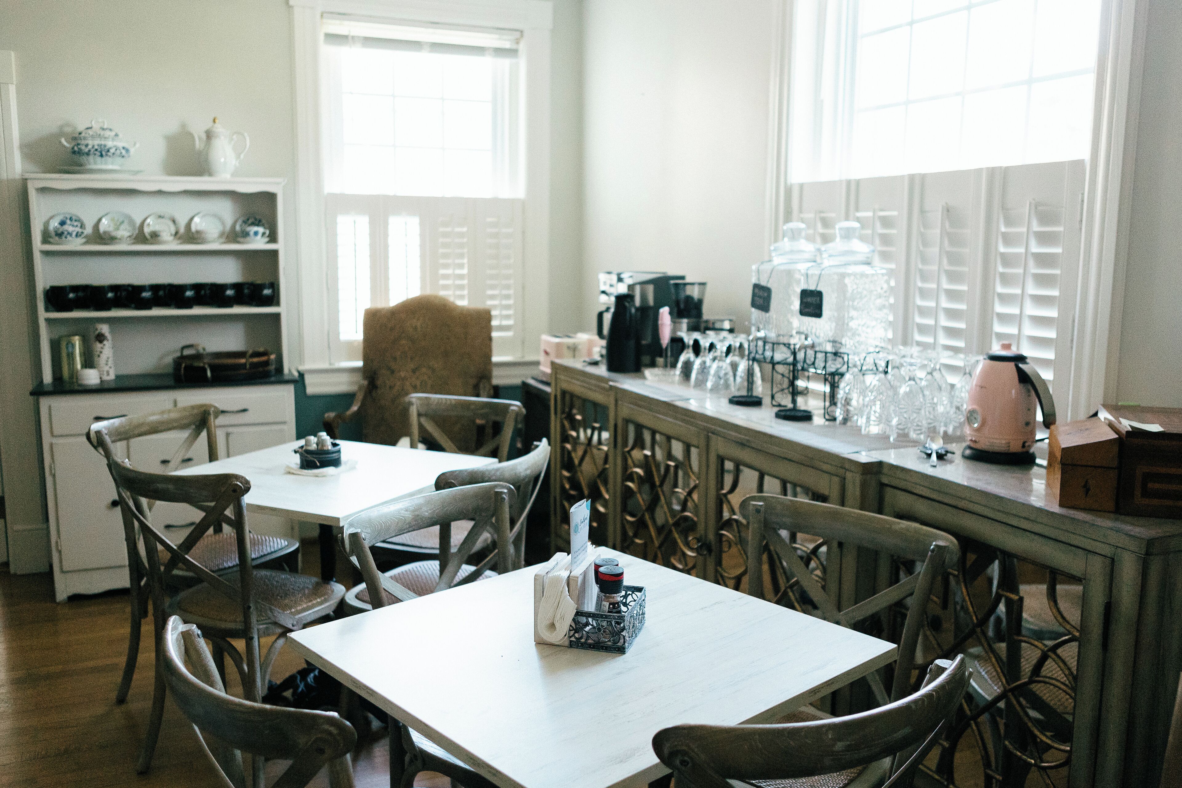 A dining area with a table and chairs, a shelf with dishes, and a window with blinds.