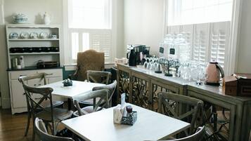 A dining area with a table and chairs, a shelf with dishes, and a window with blinds.