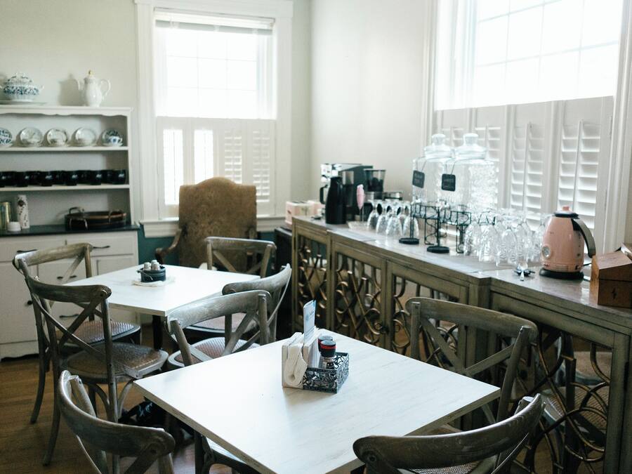 A dining area with a table and chairs, a shelf with dishes, and a window with blinds.