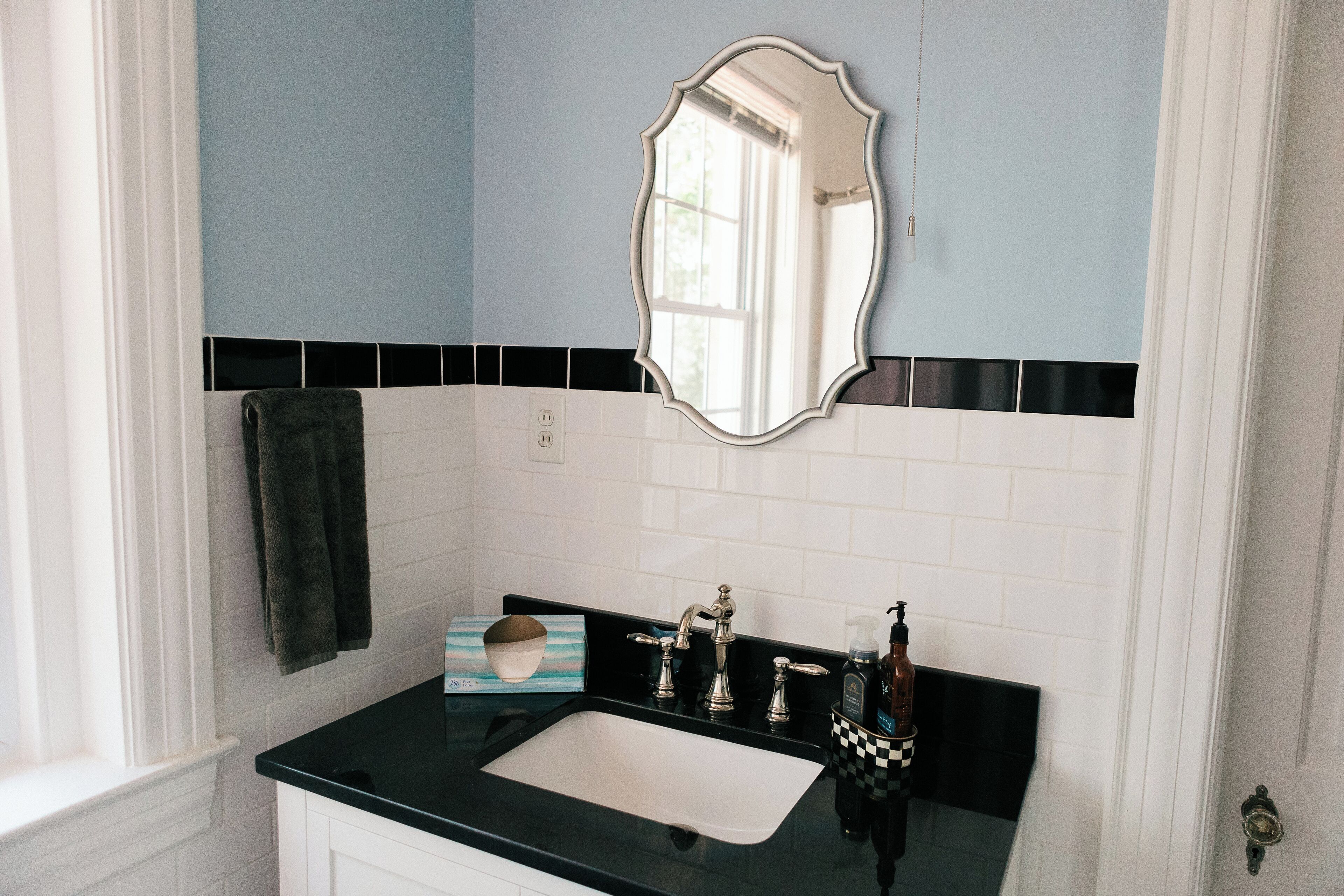 Un baño con encimera negra, azulejos blancos, un espejo, una toalla y un dispensador de jabón.