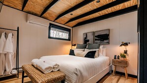 A bedroom with a wooden ceiling, a bed with white bedding, a bedside table with a lamp, and a window with a view of trees.