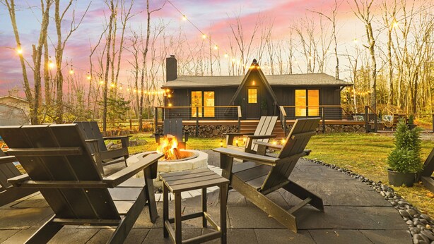 A cozy outdoor seating area with Adirondack chairs and a fire pit, surrounded by a stone wall and a view of a cabin.