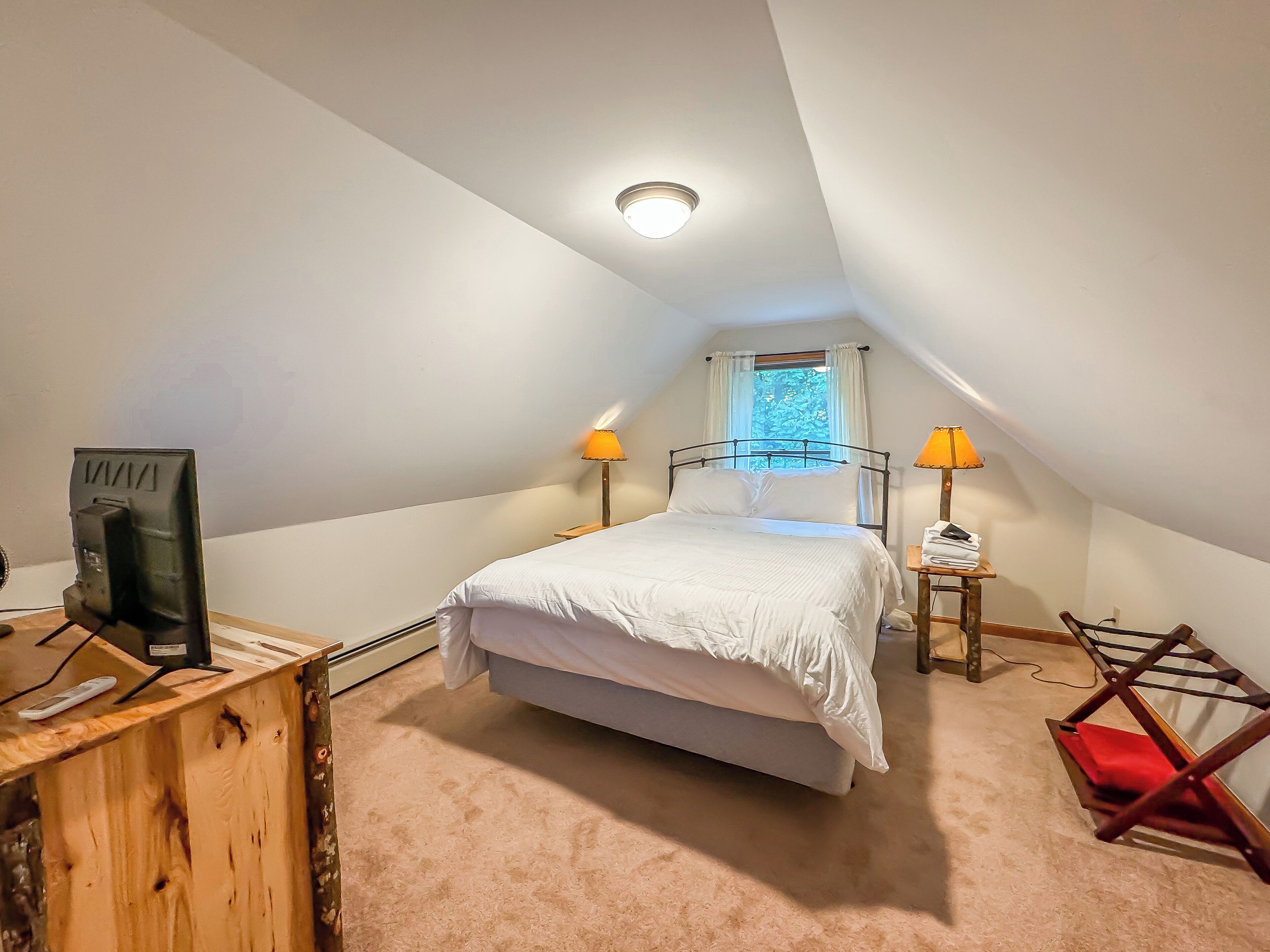 Une chambre à coucher avec un lit, deux lampes de chevet, un bureau en bois avec un ordinateur, une chaise rouge et une fenêtre donnant sur des arbres.