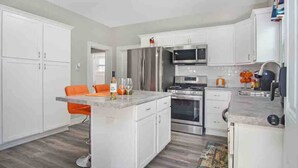 A modern kitchen with white cabinets, a central island, stainless steel appliances, and orange chairs. - Monthly Stays at Brightside Bungalow: Near Downtown Muskegon (Muskegon)