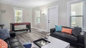 A living room with a pool table, a black leather couch with colorful cushions, a coffee table, and a door leading to another room. - Monthly Stays at Brightside Bungalow: Near Downtown Muskegon (Muskegon)