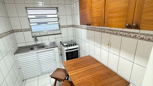 Une cuisine de taille modeste avec des murs recouverts de carreaux blancs, une table en bois avec des bancs, une cuisinière à gaz et une fenêtre avec des stores.