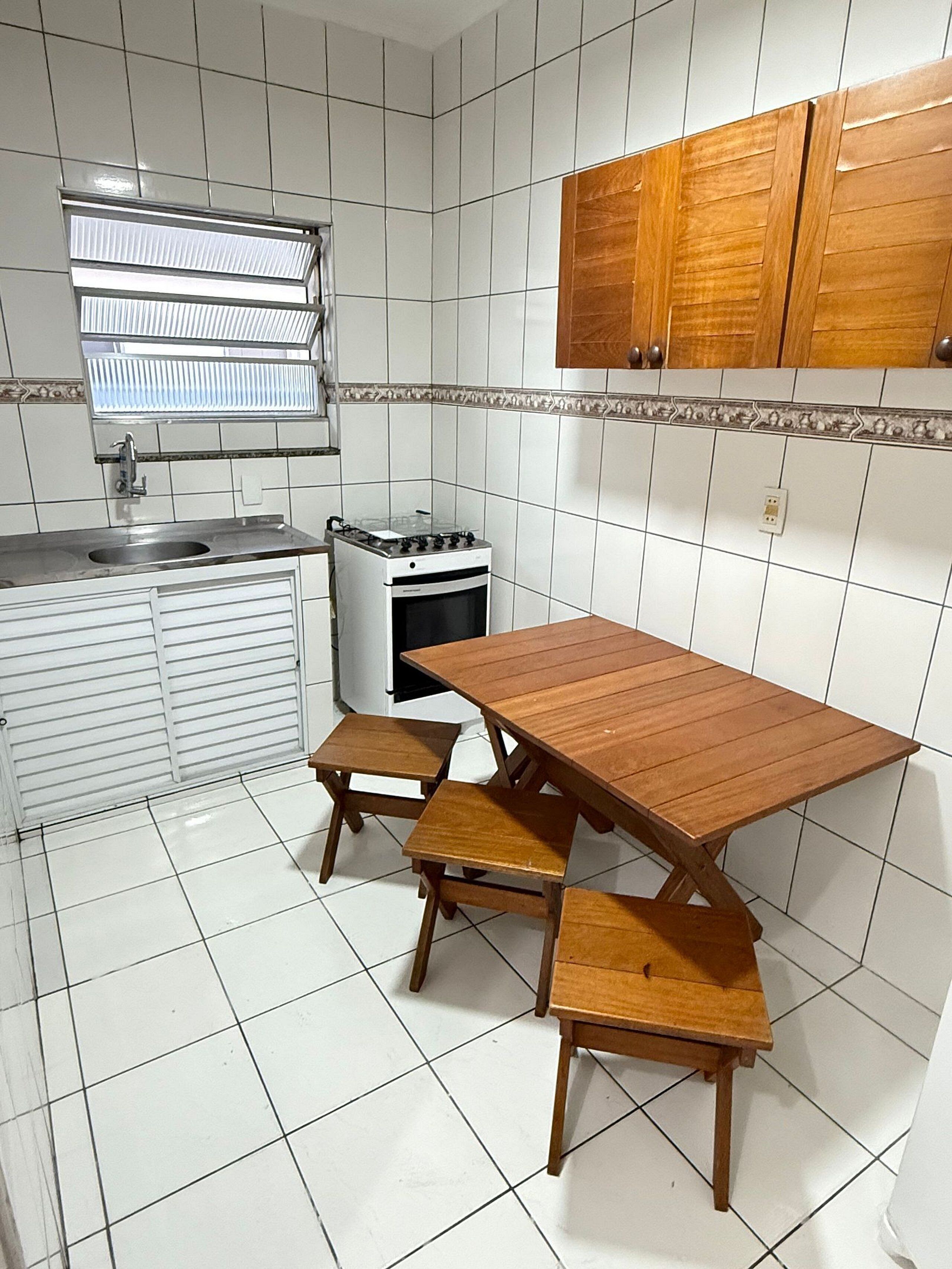 A compact kitchen with white tiled walls, a wooden table with benches, a gas stove, and a window with blinds.