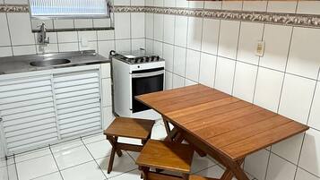A compact kitchen with white tiled walls, a wooden table with benches, a gas stove, and a window with blinds.