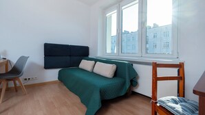 A small, neatly arranged bedroom with a single bed, a chair, a desk, and a window with a city view. - Uniwersytecka Standard Studio (Lódz)