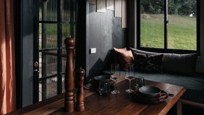 Un coin repas avec une table en bois dressée pour quatre personnes, une fenêtre donnant sur un champ verdoyant et une porte noire avec un panneau en verre.