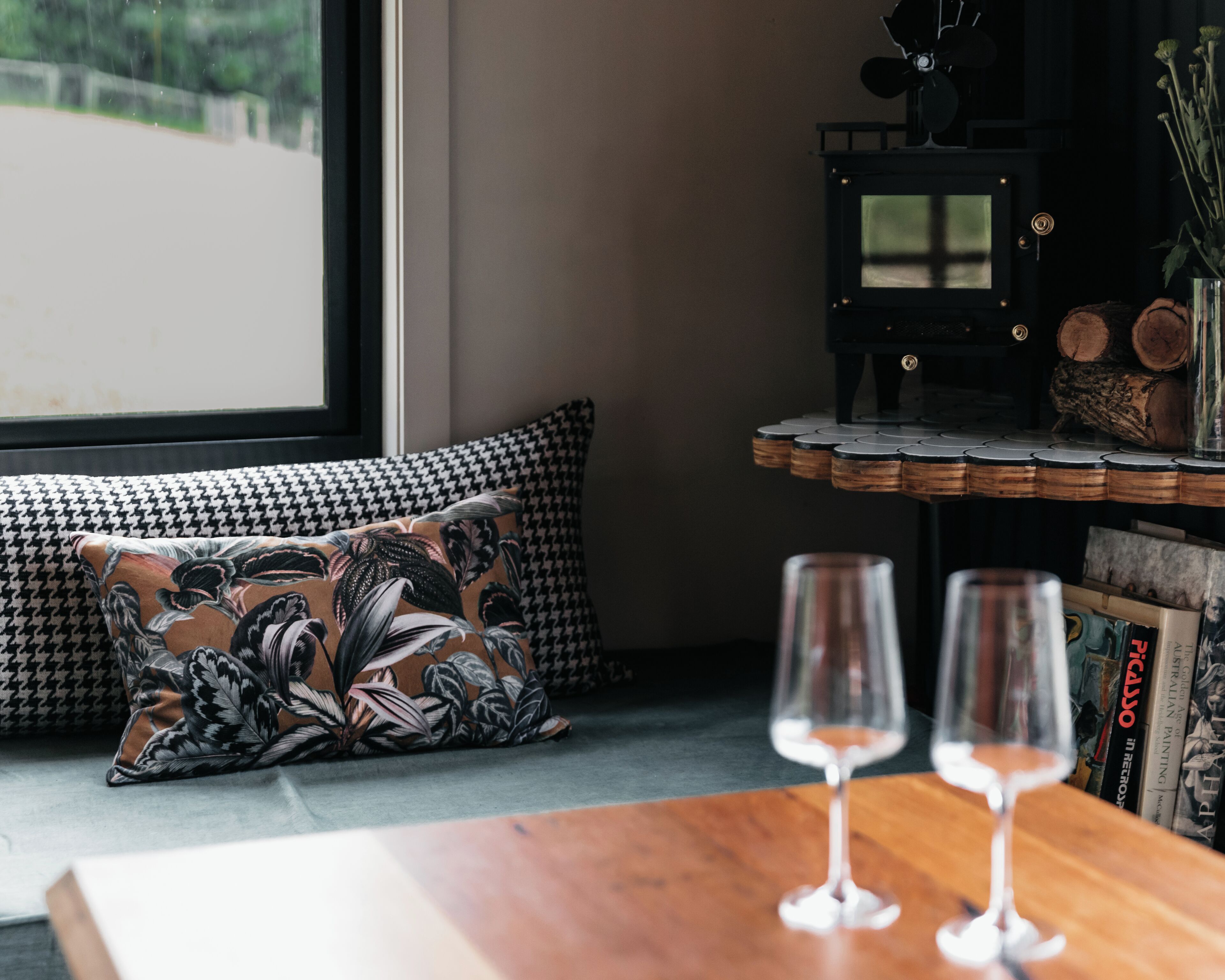 A cozy seating area with a patterned pillow, a small table with two wine glasses, and a bookshelf with books and decorative items.