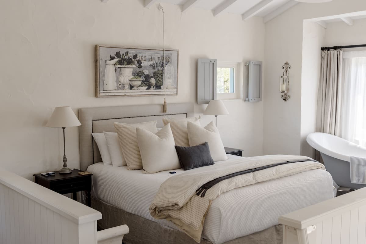 A bedroom with a bed, bedside tables, a nightstand, a wall-mounted lamp, a mirror, a window with curtains, and a bathtub.