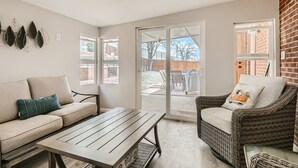 Una sala de estar con un sofá, una silla de mimbre y una mesa. Hay ventanas y vistas al exterior.