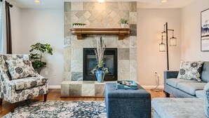 Una sala de estar con chimenea de piedra, dos sillones estampados, un otoman azul y un sofá azul.