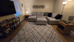 Una sala de estar con un sofá modular, un televisor de pantalla plana, una mesita de madera y un tapete estampado.