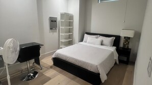 Un dormitorio con cama, una mesita de noche, lámpara, ventilador y estantería.