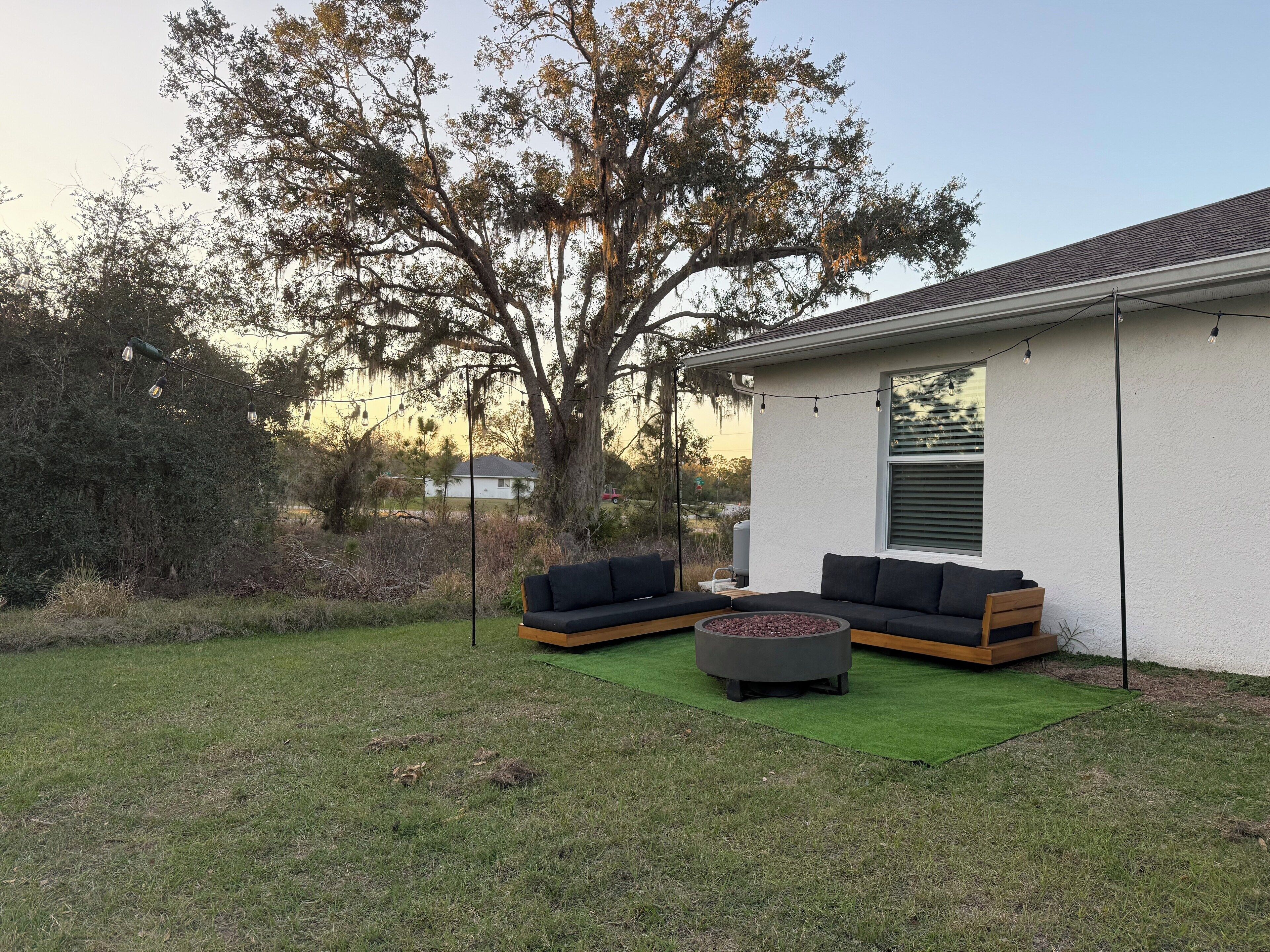 A patio with two sofas and a round table, surrounded by a lawn and a white building with a window.