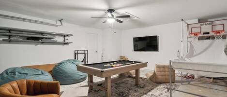 Een kamer met een pooltafel, een plafondventilator, een basketbalring en een televisie.