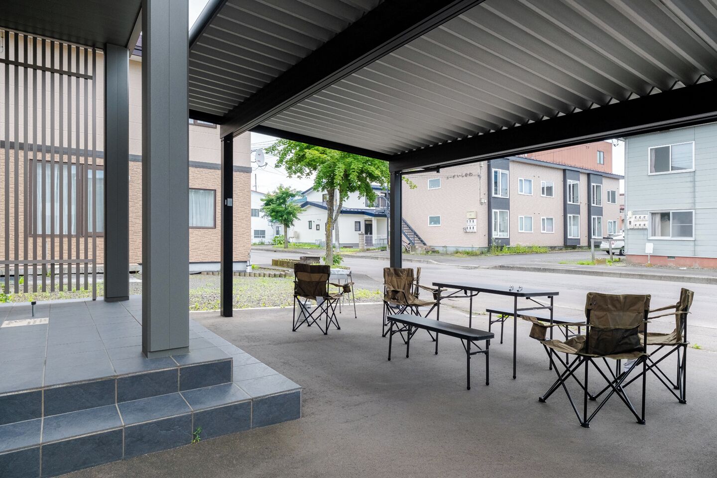 An outdoor covered seating area with foldable chairs and tables, a staircase leading to an upper level, and a view of residential buildings in the background.