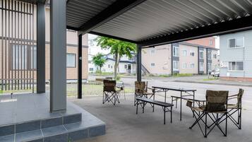 An outdoor covered seating area with foldable chairs and tables, a staircase leading to an upper level, and a view of residential buildings in the background.