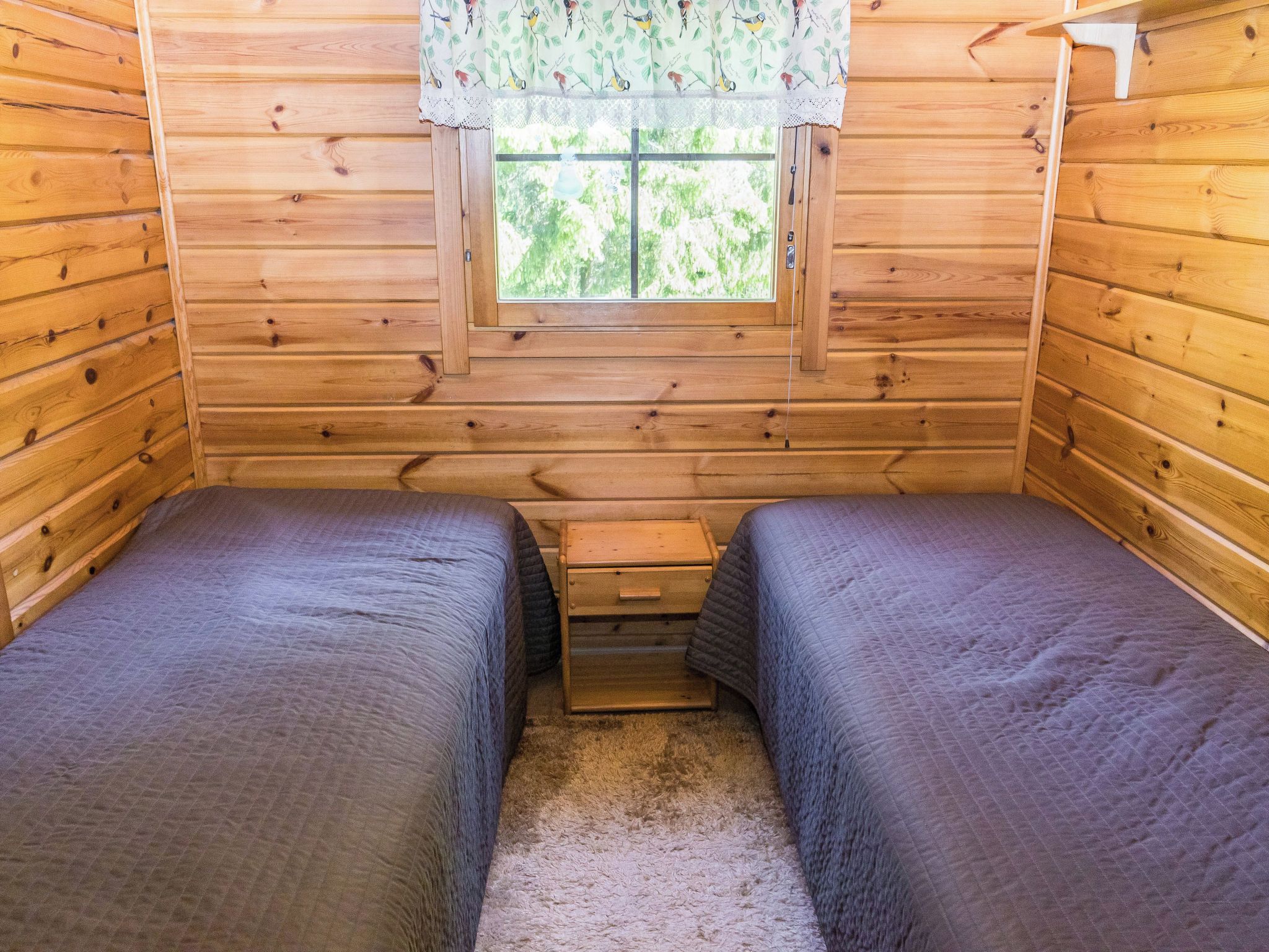 A cabin room with two beds, a window with a lace curtain, and a small wooden nightstand.