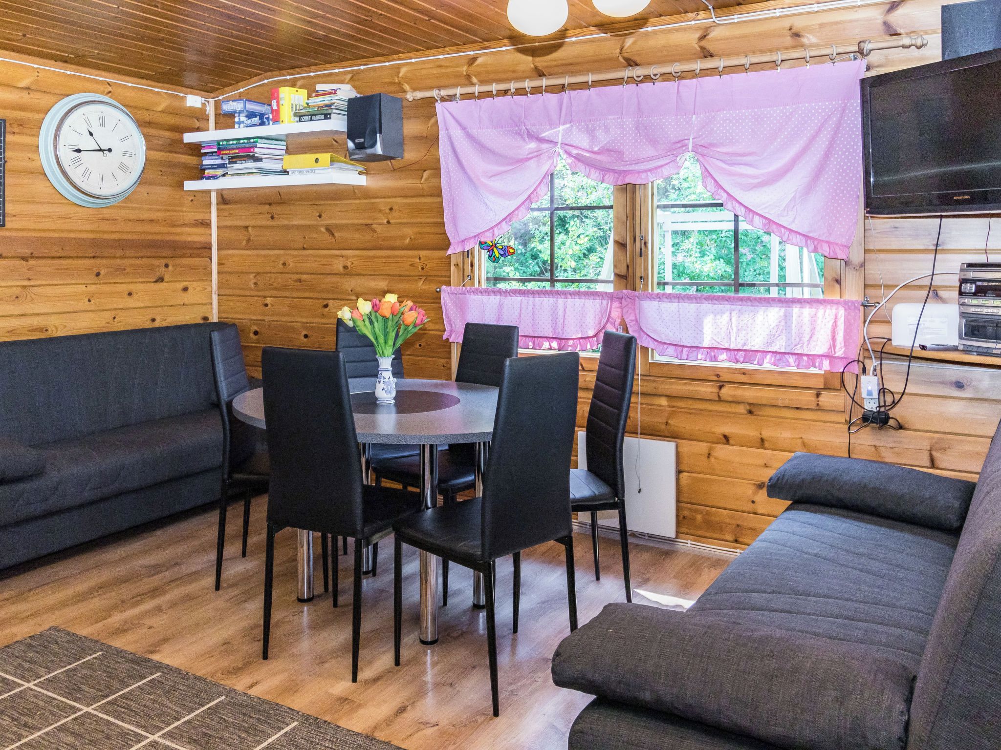 Een knus chaletinterieur met een eettafel, stoelen, een bank, een klok en een raam met gordijnen.