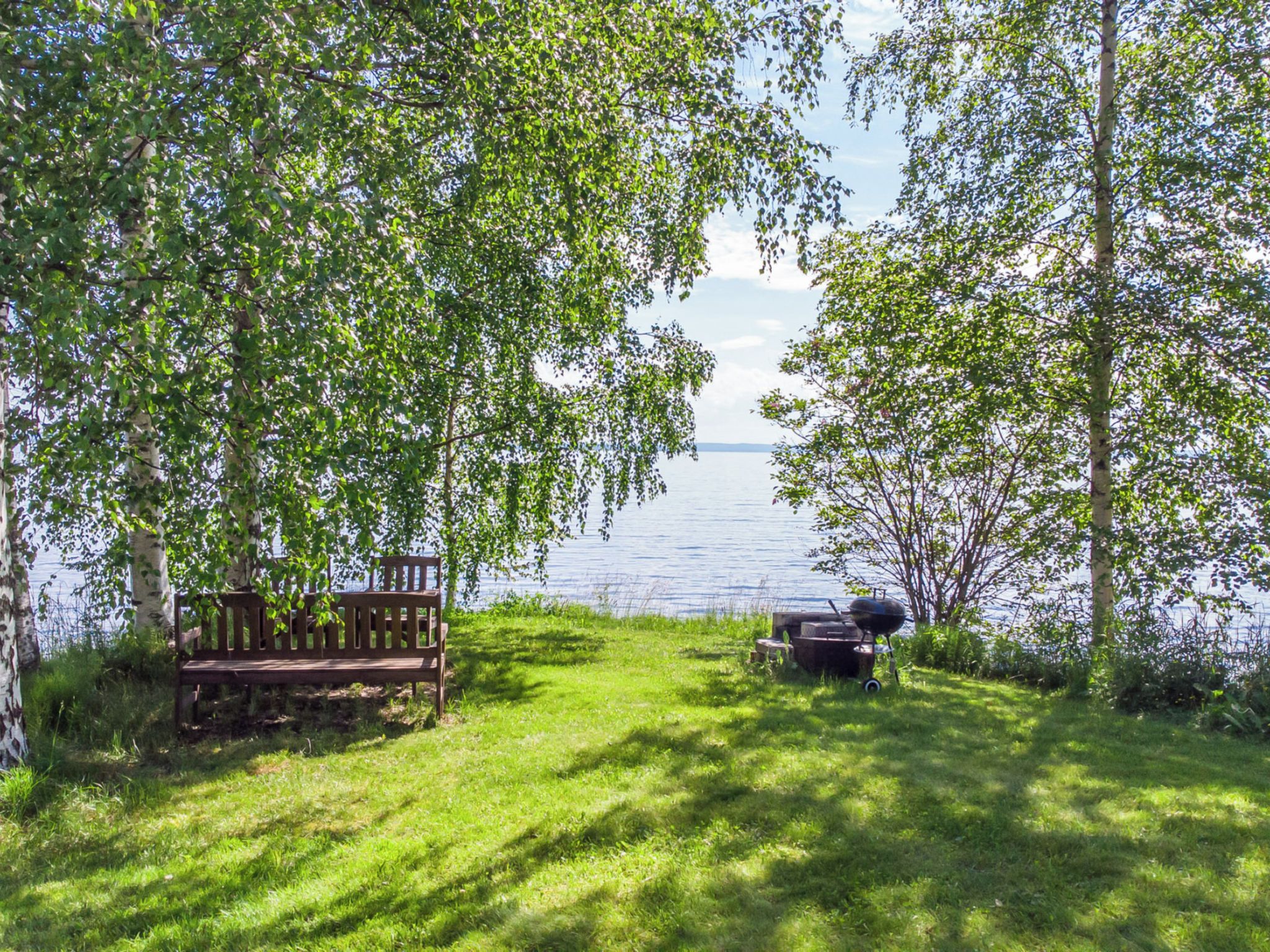 Een gazon met een houten bank en een barbecue, omringd door bomen en met uitzicht op een waterpartij.