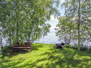 Een gazon met een houten bank en een barbecue, omringd door bomen en met uitzicht op een waterpartij.