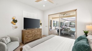 Un dormitorio con cama, televisor, cómoda de madera, sofá gris y vistas a una piscina y zona de estar al aire libre.