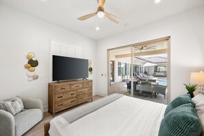 A bedroom with a bed, a TV, a wooden dresser, a grey sofa, and a view of a pool and outdoor seating area.