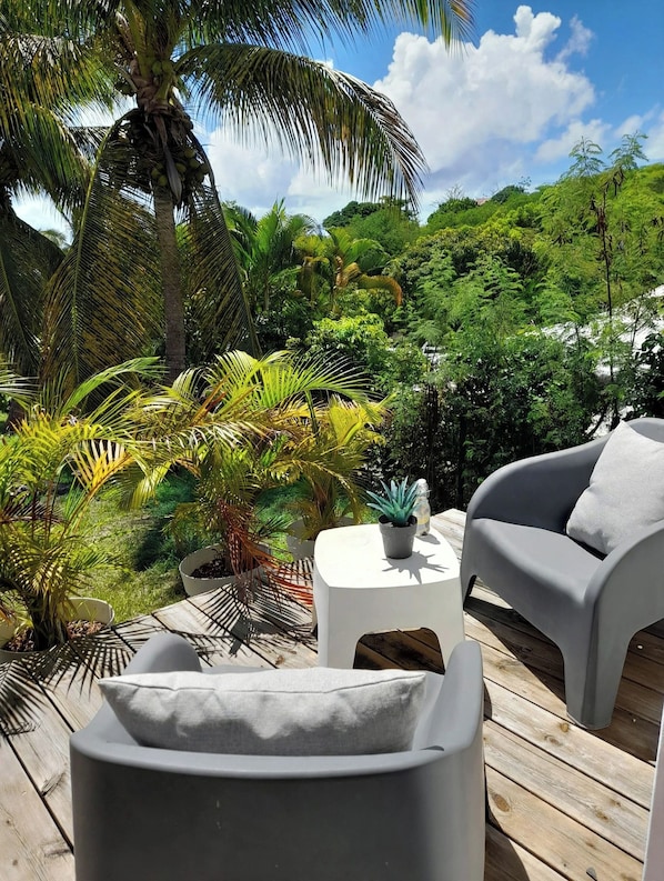 A wooden deck with a grey sofa and a small table, surrounded by lush tropical plants and a palm tree.