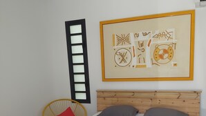 Ein Schlafzimmer mit einem hölzernen Bett, einem kleinen runden Tisch, einem gelben Stuhl und einem großen abstrakten Gemälde an der Wand.