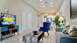 A modern living room with a flat-screen TV, a blue armchair, a sofa, a coffee table, and a dining area in the background. - SWFL: Lake McGregor 3 bedroom Home - Entire Home! (Fort Myers)