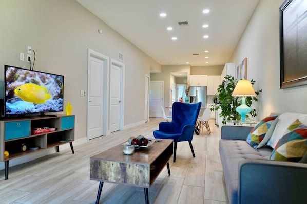 A modern living room with a flat-screen TV, a blue armchair, a sofa, a coffee table, and a dining area in the background.