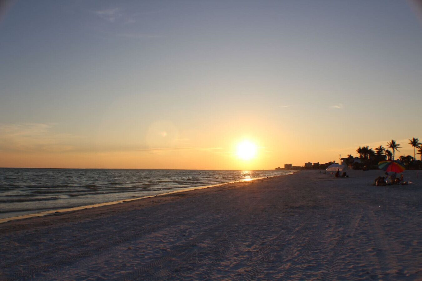 Una playa con un atardecer, palmeras y algunas personas.