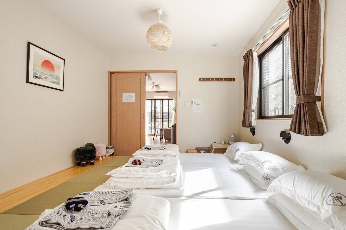 A hotel room with two beds, a large window with curtains, a hanging light fixture, and a framed picture on the wall.