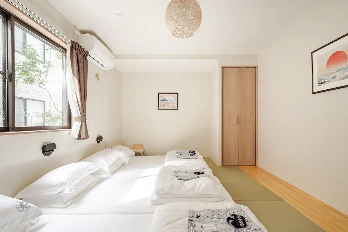 A hotel room with a bed, a bedside table, a window with curtains, an air conditioning unit, a framed picture on the wall, and a wooden door.
