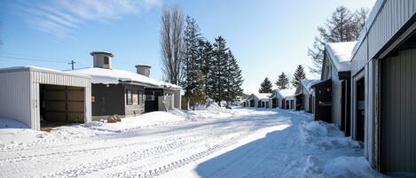 A snow-covered street with buildings on either side, including a garage and a building with a large tree in front.