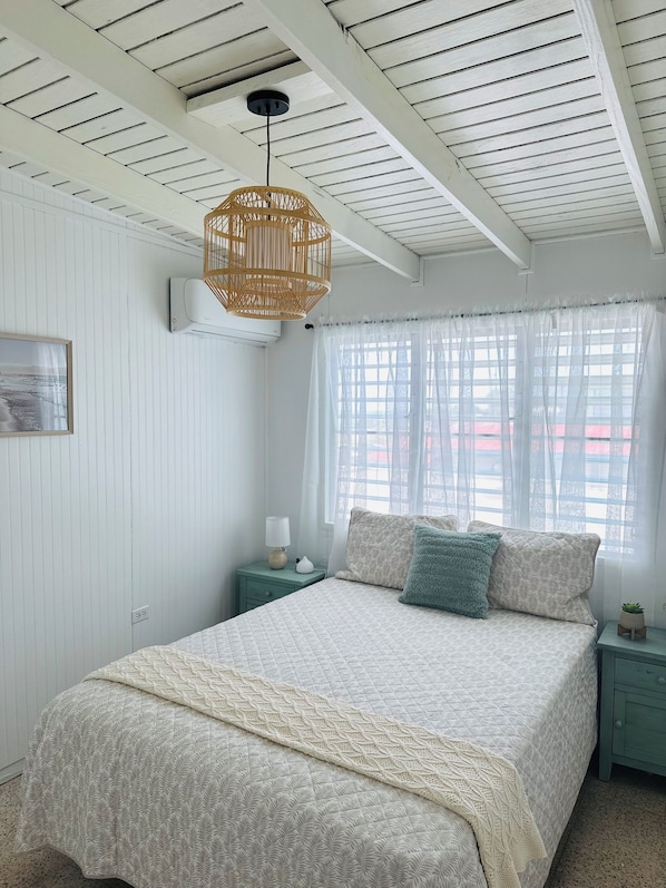 A bedroom with a bed, a bedside table with a lamp, a hanging light fixture, and a window with sheer curtains.