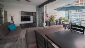 Una sala moderna con televisión de pantalla plana, sofá, mesa de comedor y balcón con plantas y una sombrilla.