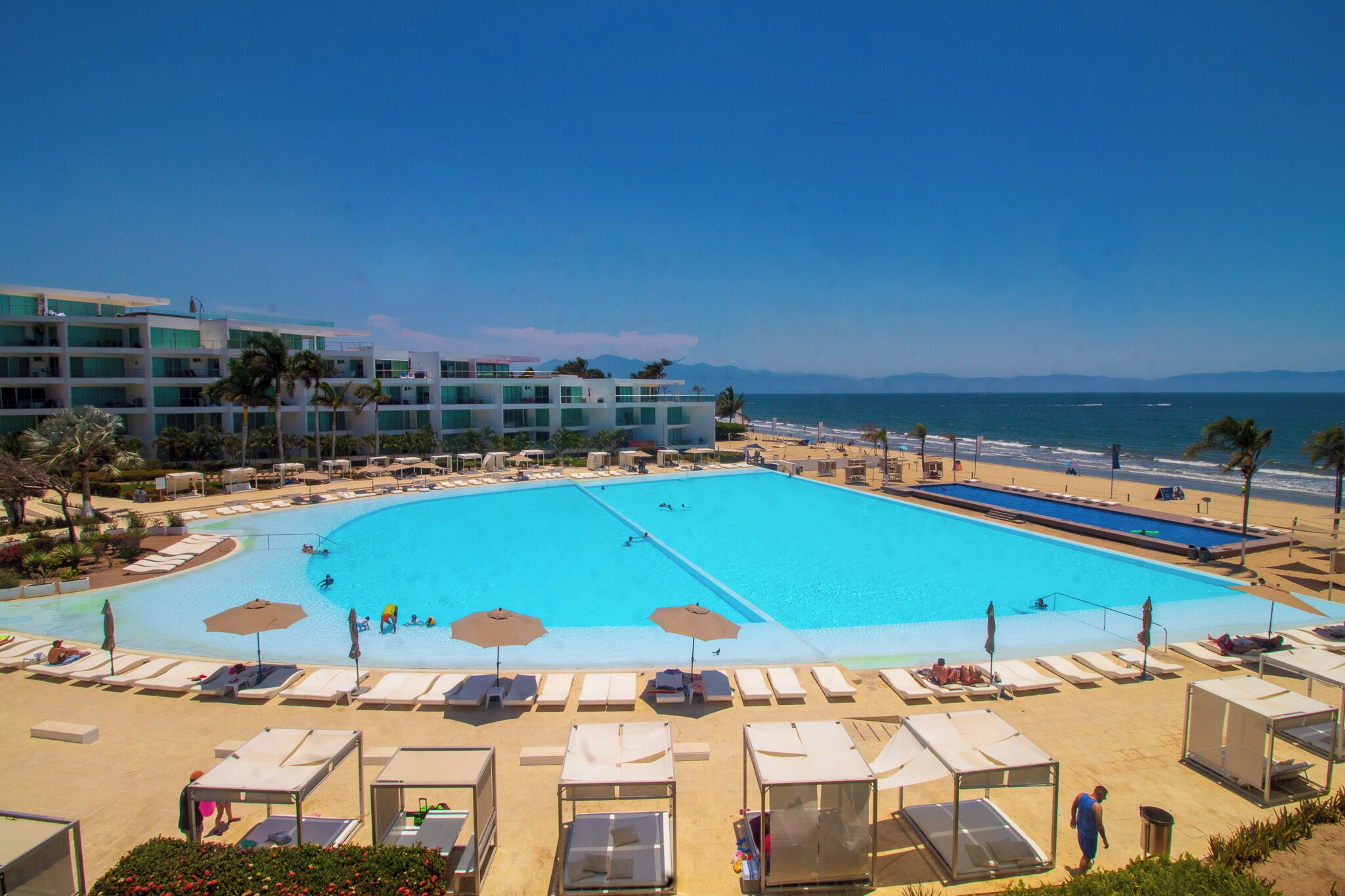 Ein großer Pool mit Liegestühlen und Sonnenschirmen, umgeben von einem mehrstöckigen Gebäude und einem Strand im Hintergrund.